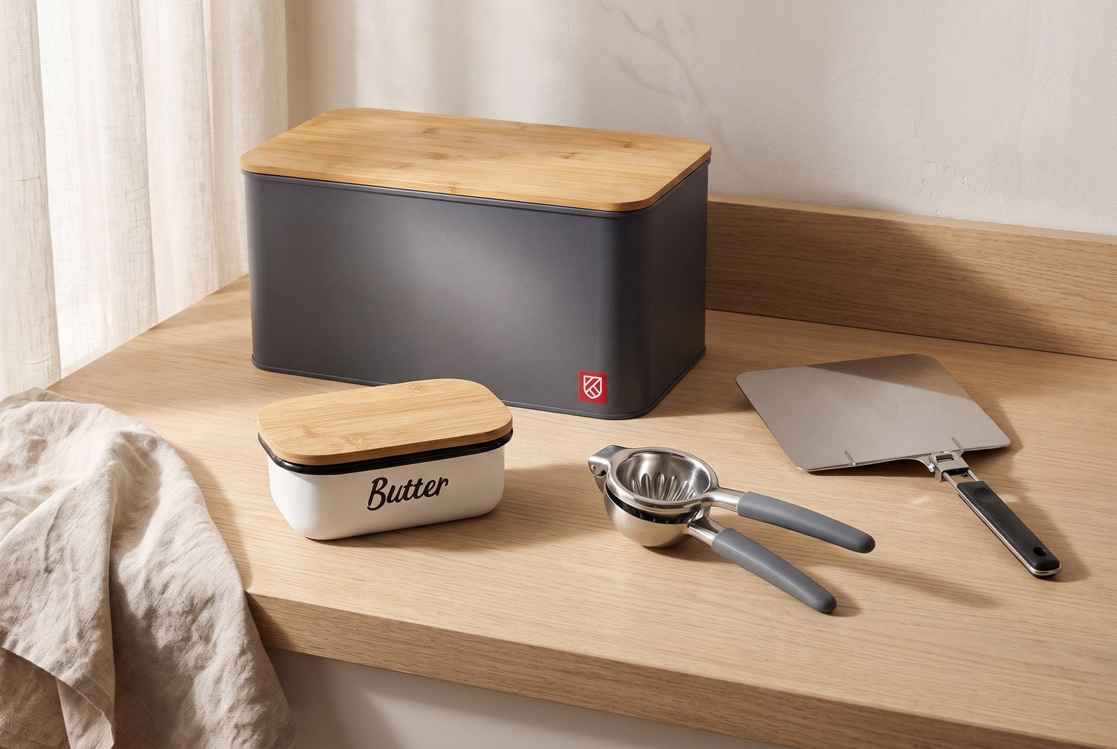 Bread box, butter dish, lemon squeezer and folding pizza peel on an oak worktop beside a window